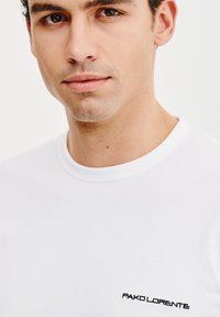 Biała bawełniana koszulka z okrągłym dekoltem i krótkimi rękawami. Posiada czarny haftowany tekst po lewej stronie z nazwą marki.