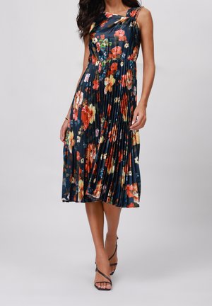 Kobieta w granatowej plisowanej sukience midi bez rękawów z wielokolorowym kwiatowym wzorem, w połączeniu z czarnymi sandałkami na paski.