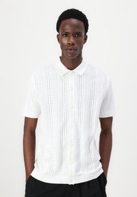 Baltā īsām piedurknēm adītā polo krekls ar reljefu rakstu, pogu priekšpusi un ribota apkakle un apakšmalu.