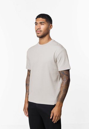 Jeune homme aux cheveux courts et tatouages sur les deux bras, portant une chemise beige à manches courtes et un pantalon noir, debout devant un fond blanc.