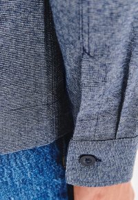 Zbliżenie na niebieską, teksturowaną mankietę koszuli z czarnym guzikiem, noszoną na niebieskich dżinsach.