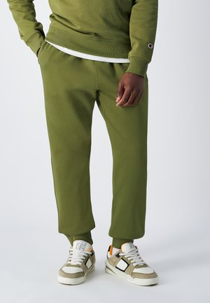 Pantalons de jogging vert olive en mélange de coton doux, dotés d'une taille élastique et de chevilles resserrées, associés à des baskets beiges et blanches.