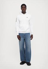 Sweatshirt branca com bolso frontal, capuz ajustável com cordão e detalhes em preto, combinado com jeans de perna larga azul clara e sapatos pretos.