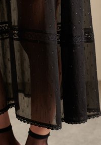 Czarna przezroczysta sukienka z pionowymi fałdami, ozdobiona małymi cekinami i koronkowym wykończeniem na dolnym brzegu oraz w pasie. Podkreśla elegancję.