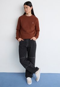 Hanorac maro, pantaloni largi negri cu cusături în contrast și adidași bej. Texturi moi, design minimalist și buzunare bine definite.