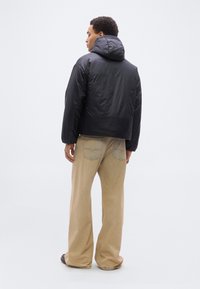 Άνδρας που φοράει μαύρο μπουφάν με κουκούλα και φαρδιά μπεζ παντελόνια, στέκεται γυρισμένος με την πλάτη σε απλό ανοιχτόχρωμο φόντο.