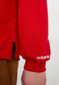 Red athletic jacket with zippered pockets, ribbed cuffs, and white Adidas logo. Made of smooth fabric with a fitted design.