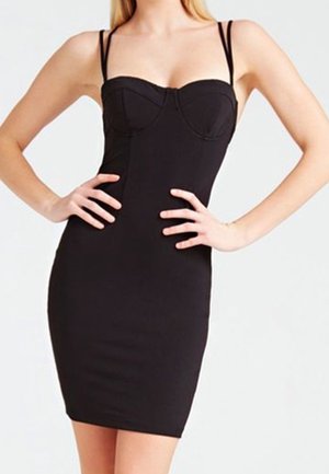 Kvinde iført en tætsiddende sort ærmeløs kjole med tynde stropper og struktureret kavalergang, stående med hænderne på hofterne mod en lys baggrund.