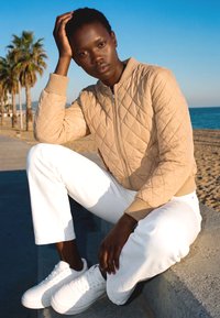 Person trägt eine beige gesteppte Jacke, weiße Hose und weiße Turnschuhe und sitzt auf einer Betonmauer am Strand mit Palmen und klarem Himmel.