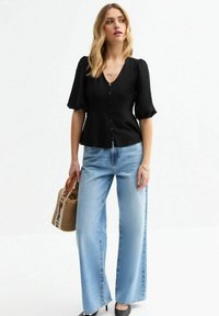 Blusa negra texturizada con escote en V y botones al frente, combinada con pantalones vaqueros anchos de color azul claro y tacones negros. Lleva un bolso tejido en la mano.