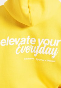 Felpa gialla con cappuccio, con intervalli di testo bianco che recita "eleva il tuo quotidiano" e "Smilodox - Lo sport è uno stile di vita." Tessuto morbido.