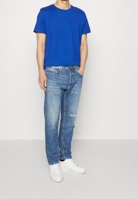 Camiseta de algodón azul con mangas cortas y un pequeño logo en el pecho, combinada con jeans de mezclilla azul claro y zapatillas blancas.