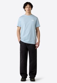 Camiseta de algodón azul claro con cuello redondo y mangas cortas, que presenta un logo blanco en el lado izquierdo del pecho, combinada con pantalones cargo negros.