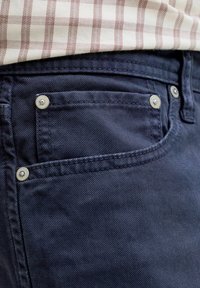 Κοντινή λήψη τσέπης σε σκούρο μπλε τζιν με ασημένια καρφιά, φορεμένη με μπεζ και κόκκινο καρό πουκάμισο.