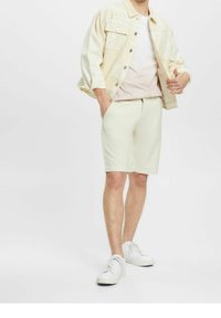 Giacca di jeans beige chiaro con due tasche sul petto, abbinata a una t-shirt pastello e pantaloni corti chiari. Sneakers bianche completano l'outfit.