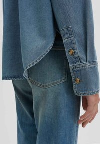 Camicia di jeans di colore azzurro chiaro, con orlo appuntito, polsini con bottone e dettagli di cucitura in evidenza, abbinata a jeans coordinati.