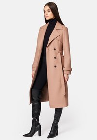 Beżowy płaszcz typu trench z wełny, z podwójnym zapięciem na guziki, szerokimi klapami, kieszeniami po bokach i paskiem w talii. Stylizacja z dopasowaną czarną bluzką i kozakami do kolan.