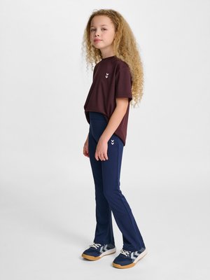 Jong meisje met krullend blond haar, draagt een bordeauxrood shirt met korte mouwen, donkerblauwe broek en donkerblauwe sneakers met witte veters, staand opzij.