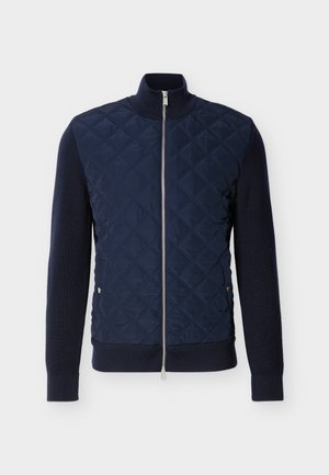 Veste matelassée bleu marine avec fermeture éclair intégrale à l'avant, col et poignets côtelés, et poches latérales à boutons-pression sur fond uni.