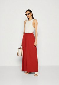 Bež brezrokavnik kombiniran z rdečo maxi krilo z robom. Krilo ima nabran pas in je izdelano iz gladkega blaga. Dodatki: majhna ročna torbica.