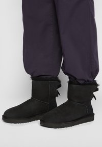 Des bottines noires en suède présentent une doublure en fourrure douce, un bout rond et des accents décoratifs sur le côté. Elles sont associées à un pantalon violet ample.