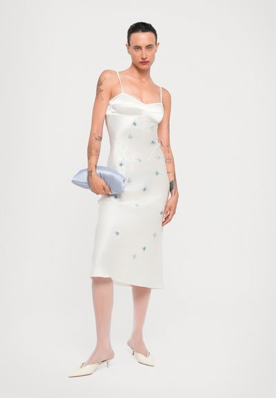Satin wit jurkje met dunne bandjes en een aansluitende pasvorm, voorzien van blauwe bloemenborduring. Model houdt een lichtblauwe clutch tas vast.