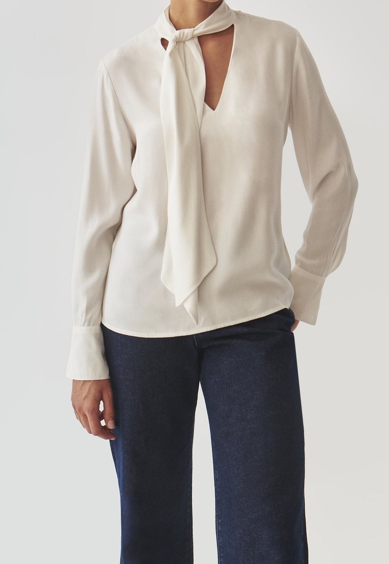 Femme portant un chemisier crème à manches longues avec un foulard noué autour du cou et un jean bleu foncé, debout devant un fond uni.