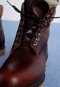 Stivale in pelle marrone con lacci neri e occhielli in argento, superficie texturizzata, dettagli in tono naturale e una robusta suola in gomma.