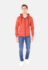 Felpa zip arancio ruggine con cappuccio regolabile, tasche frontali e polsini a costine, abbinata a jeans blu strappati e sneakers bianche.