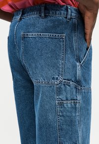 Άτομο που φοράει μπλε τζιν παντελόνι με μεγάλες πίσω και πλαϊνές τσέπες, το χέρι ακουμπά στην πλαϊνή τσέπη, φοράει ροζ και κόκκινη ριγέ μπλούζα.