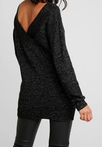 Nainen, joka on pukeutunut mustaan, kimaltavaan neulepuseroon, jossa on syvä V-mallinen selkä ja pitkät hihat, yhdistettynä mustiin nahkahousuihin, selin katsojaan.