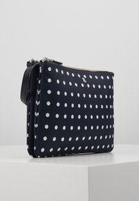 Marinblå clutchväska med prickar i en jämn textur, dragkedjestängning och avtagbar rem. Har vita prickar jämnt fördelade över ytan.