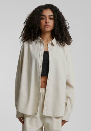 Chemise beige à boutons fabriquée en tissu doux, présentant une coupe décontractée, un col classique et des manches longues. Top noir court en dessous.