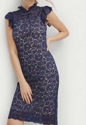 Kobieta ubrana w granatową kwiecistą koronkową sukienkę z krótkimi rękawami typu "cap" i ozdobnym, marszczonym wysokim kołnierzem związanym delikatną kokardką.