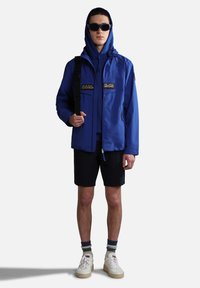 Giacca impermeabile blu con tasche frontali, abbinata a una felpa blu con cappuccio, pantaloni corti neri e sneakers bianche. Include occhiali da sole e una borsa a tracolla.