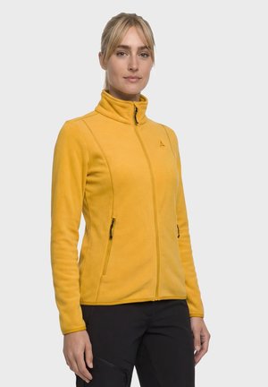 Woman wearing a yellow zip-up fleece jacket with two zipped pockets and black pants, standing against a plain light background.