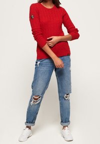 Rode gebreide trui met een textuurpatroon, ronde halslijn en logopatch op de linkermouw, gecombineerd met verwasde blauwe jeans en witte sneakers.