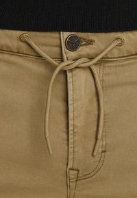 Un pantalon beige comporte une taille à cordon de serrage avec un bouton en métal brun. Le tissu est lisse avec un léger éclat et des détails de couture visibles.
