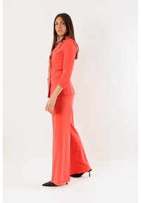 Completo corallo composto da un blazer aderente con maniche a tre quarti e pantaloni a gamba larga, abbinato a tacchi neri a punta su sfondo bianco.