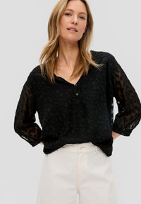 Zwarte blouse met een luipaardprint textuur, lichte doorzichtige mouwen, een V-hals en een klein knoopdetail. Gecombineerd met lichtgekleurde broeken.