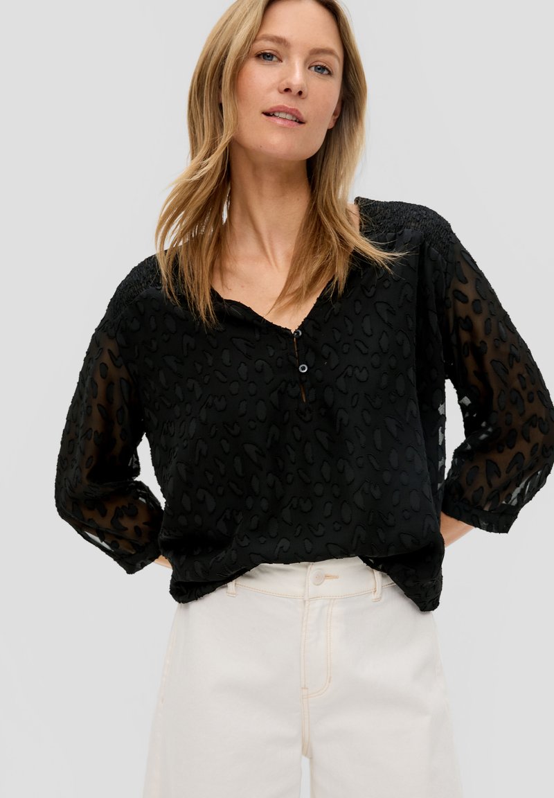 Zwarte blouse met een luipaardprint textuur, lichte doorzichtige mouwen, een V-hals en een klein knoopdetail. Gecombineerd met lichtgekleurde broeken.