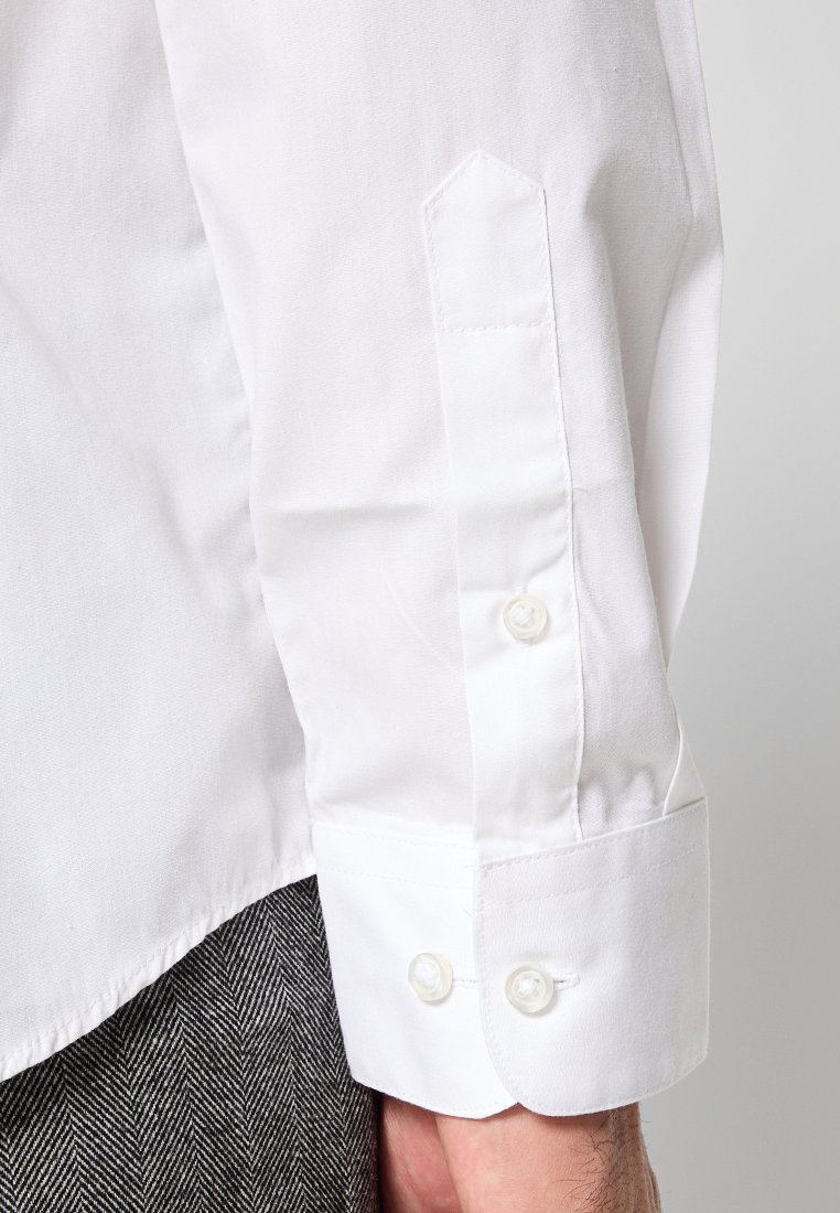 Witte katoenen overhemd met lange mouwen, voorzien van puntige manchetten, twee knoopsluitingen en een gladde textuur, gedragen over een stof met visgraatpatroon.