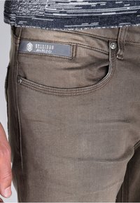 Vaqueros de mezclilla marrones con una textura suave, bolsillos frontales y un bolsillo trasero que presenta un logotipo de etiqueta gris. Incluye un botón de metal y remaches.
