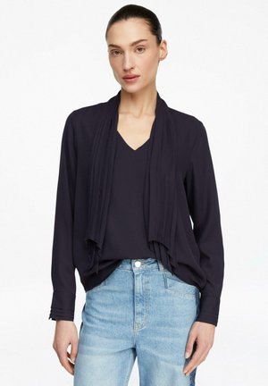 Frau trägt eine schwarze, langärmlige Bluse mit offenem Vorderteil und hellblaue Jeans mit hoher Taille vor weißem Hintergrund.