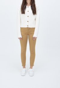 Cárdigan de punto crema con botones grandes, combinado con pantalones slim-fit marrón dorado y zapatillas blancas. Fondo neutro.