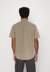 Kortärmad, ljusbrun skjorta med knäppkrage, slät textur och rundad fåll, sedd bakifrån, vilket avslöjar en ren design.