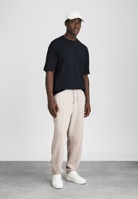 Svart kortermet t-skjorte med lomme, beige sweatpants med sidelister, og hvite sneakers, fullført med en hvit caps.