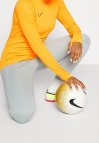 Camiseta de manga larga de Nike en naranja brillante con un cuello con cremallera, combinada con pantalones gris claro y un balón de fútbol texturizado en blanco y oro.