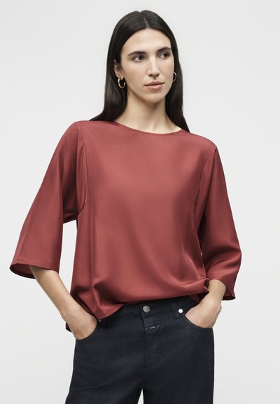 Γυναίκα με μακριά σκούρα μαλλιά που φορεί χρυσούς κρίκους στα αυτιά, μια φαρδιά, σκουρόχρωμη μπλούζα με μανίκια μέχρι τους αγκώνες και σκούρα παντελόνια με τα χέρια στις τσέπες.