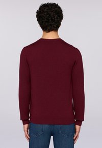 Burgundský svetr s kulatým výstřihem a ribovanými manžetami a lemem, vyrobený z měkkého materiálu, nošený s modrými džíny, viděný zezadu.